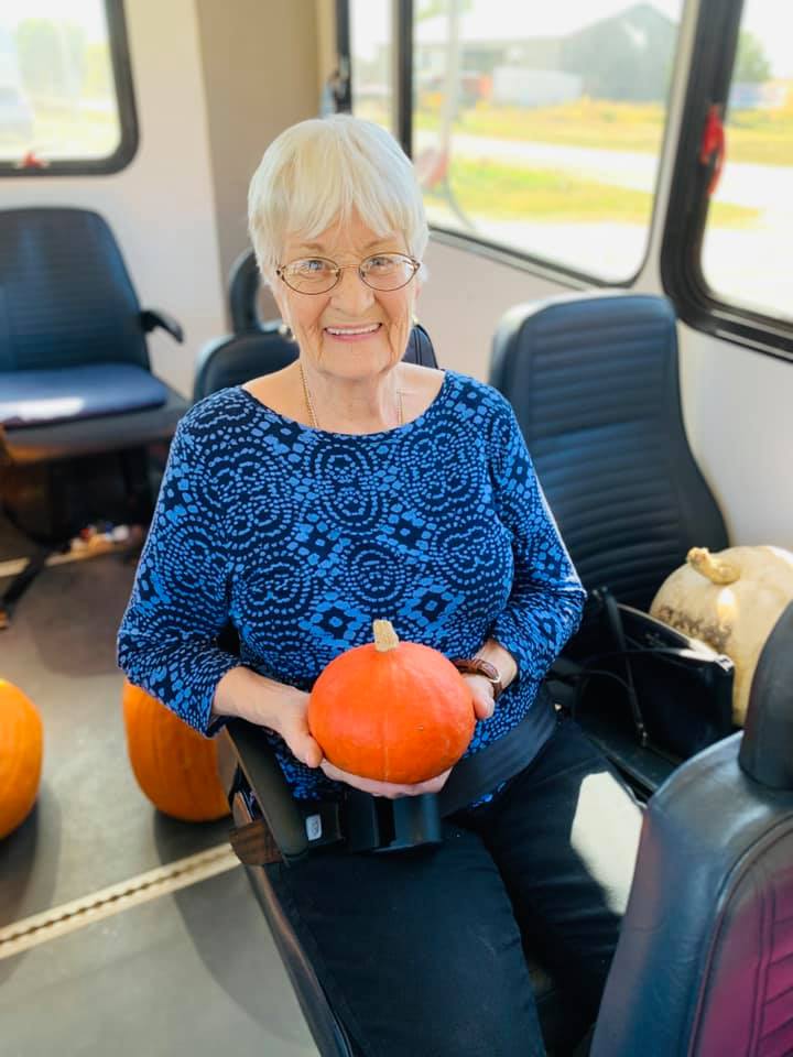 An elderly woman smiling while holding a small pumpkin inside a bus.