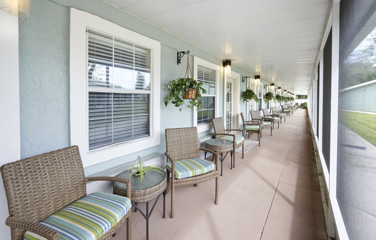 A long patio with multiple wicker chairs and small tables, adorned with hanging plants and windows along the wall.