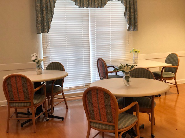 A cozy dining area with round tables and comfortable chairs, decorated with vases of flowers near a window with blinds and drapes.