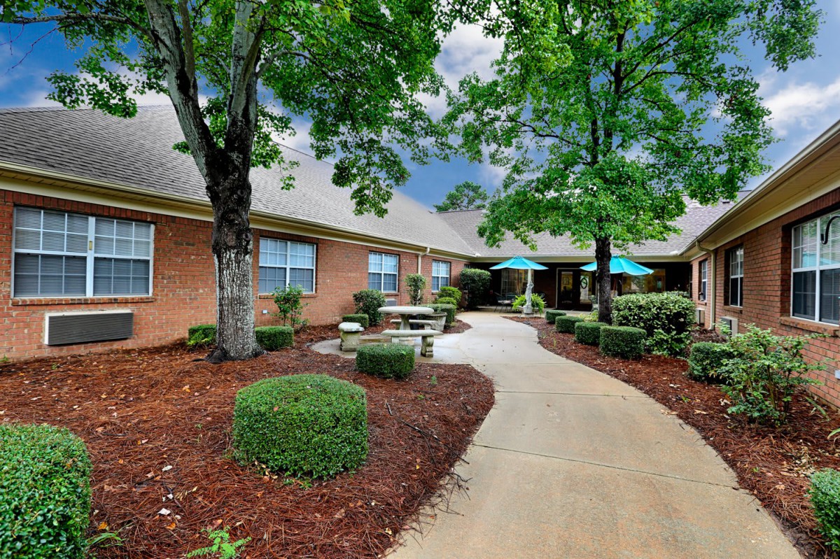 A scenic view of a well-landscaped garden path leading to a brick building, featuring outdoor seating and patio umbrellas.