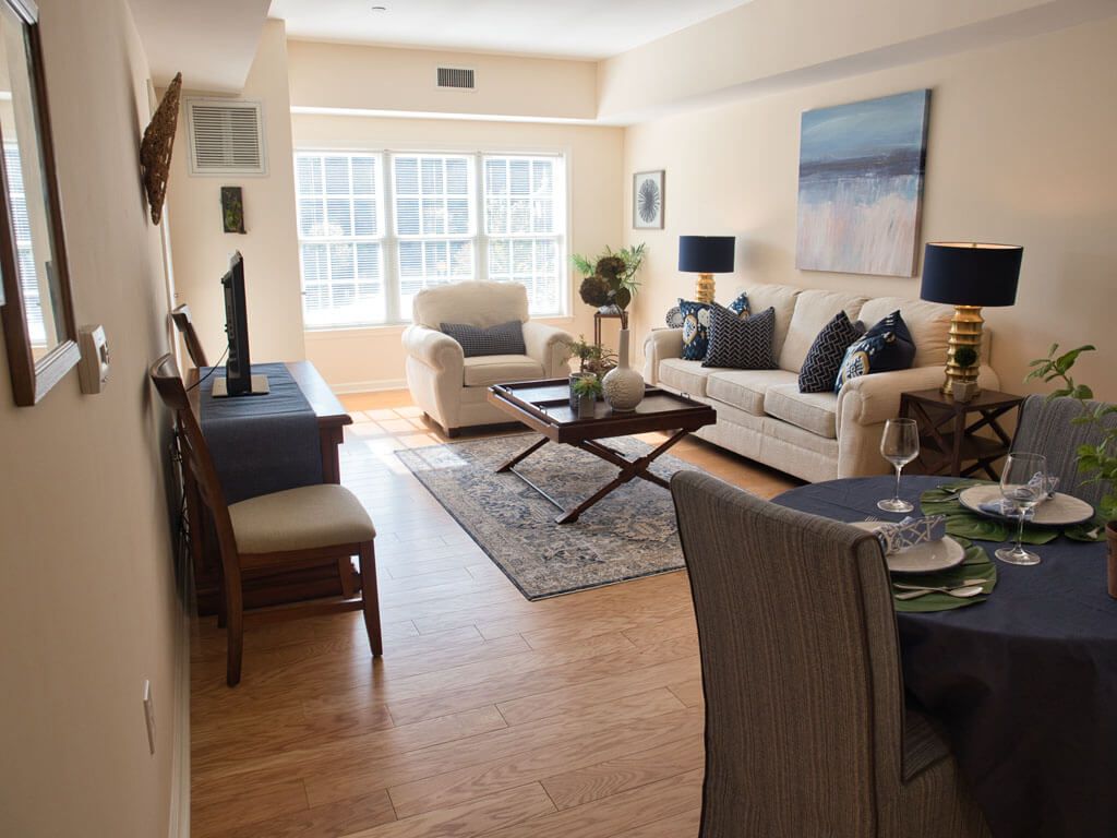 A modern living room with elegant furnishings, including a sofa, armchair, coffee table, and television. The space is decorated with artwork on the wall and a rug on the wooden floor.