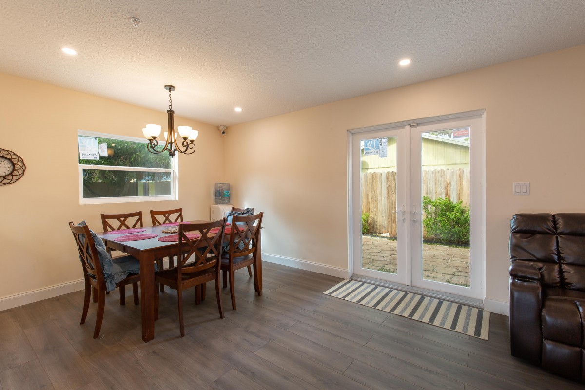 A bright dining room with a wooden table and chairs, featuring a chandelier and glass doors leading to the garden.