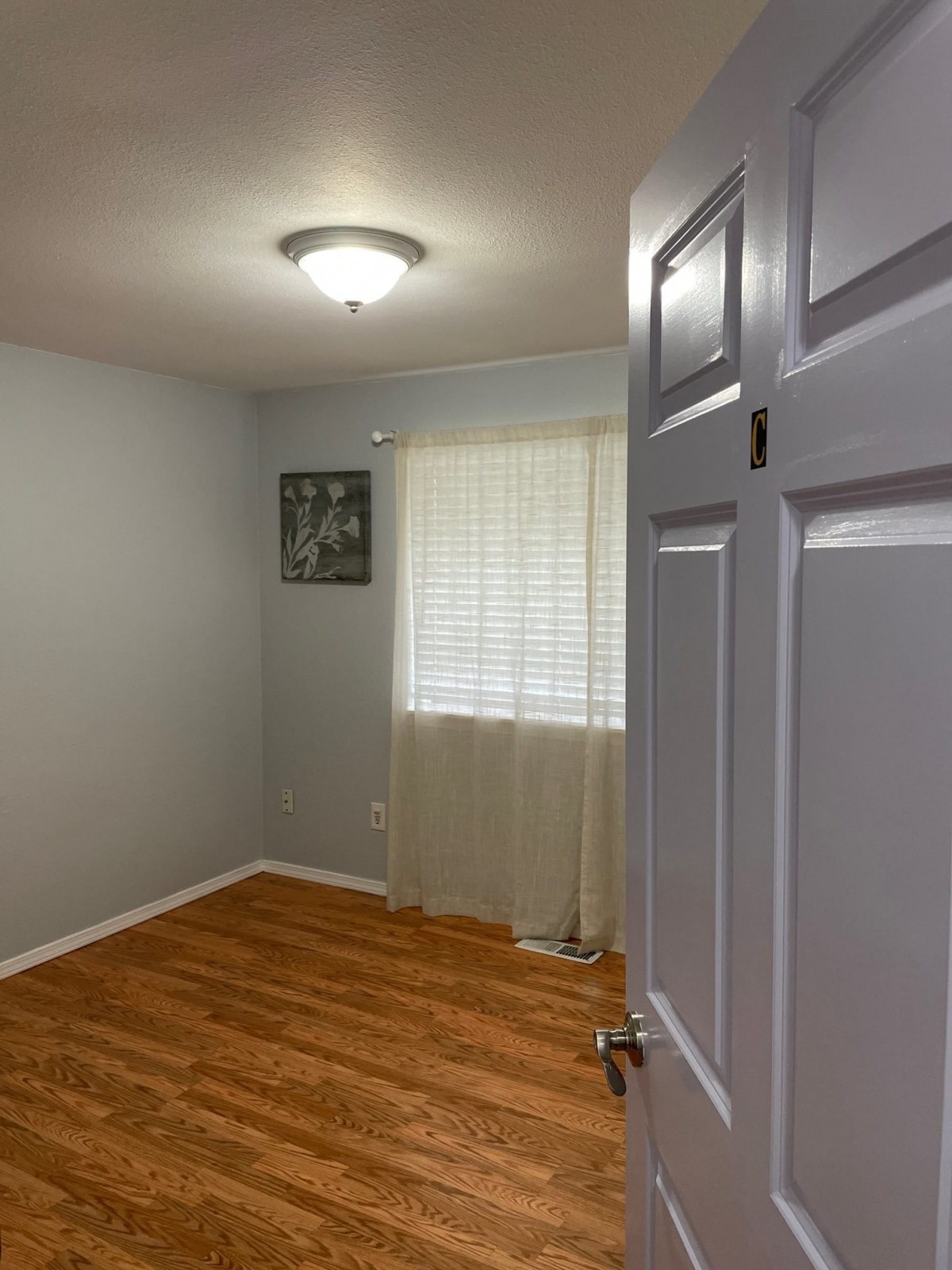 A small empty room with wooden flooring and a window with curtains.