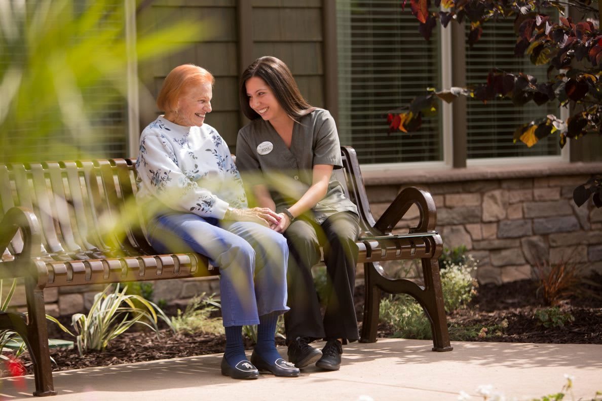 A caregiver and an elderly person share a moment on a bench outside.