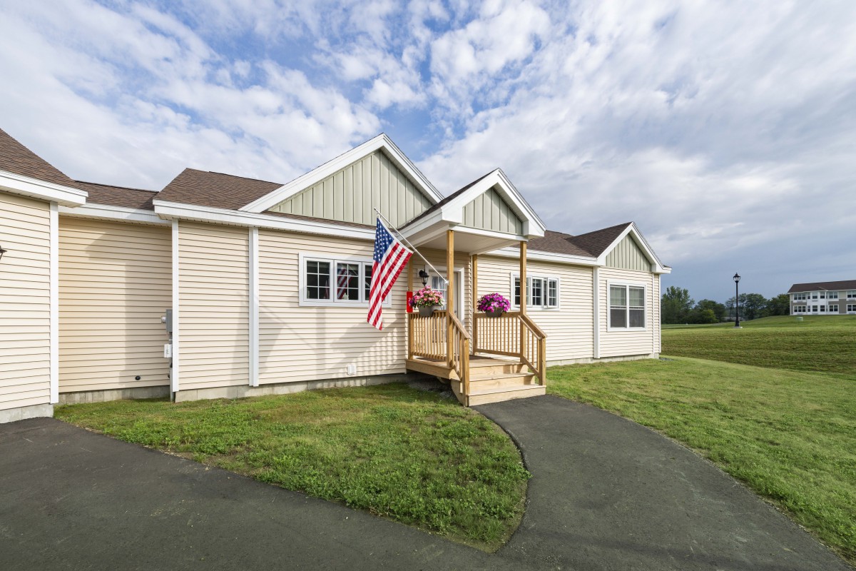 A single-story house with an American flag on the porch, set against a cloudy sky with a neatly maintained lawn.