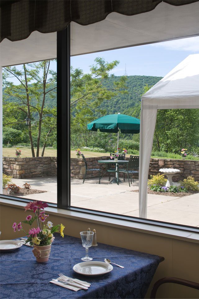 A scenic view from inside, looking out at a patio with tables and umbrellas set against a backdrop of mountains and greenery.