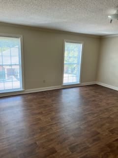 An empty room with hardwood flooring and two large windows allowing natural light to enter.