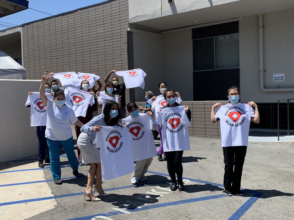 A group of people wearing masks, holding and displaying t-shirts with a logo, standing outside a building.