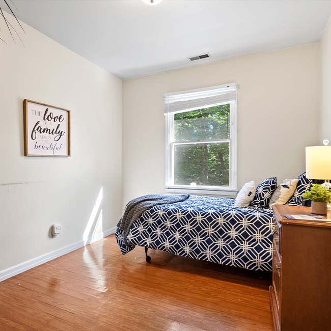 A cozy bedroom with a single bed, adorned with patterned bedding and decorative pillows, features a window allowing natural light to illuminate the hardwood floor.