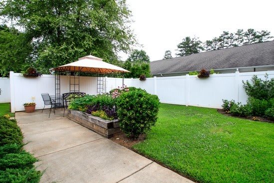 A cozy backyard featuring a patio with outdoor seating and an umbrella, surrounded by lush greenery and a white fence.