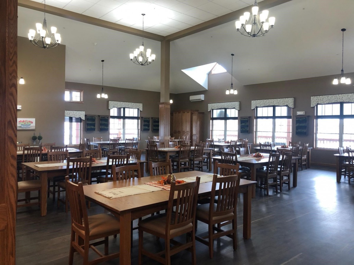 An empty dining hall with wooden tables and chairs, featuring large windows and overhead lighting.