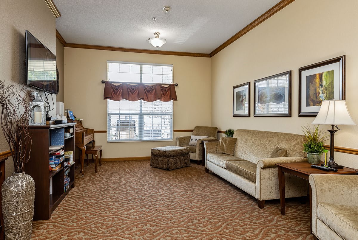 A cozy living room with a sofa, ottoman, and framed pictures on the wall, featuring a carpeted floor and a large window with curtains.