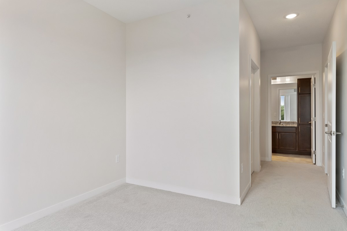 A minimalist empty room with white walls and a carpeted floor, leading to a hallway.