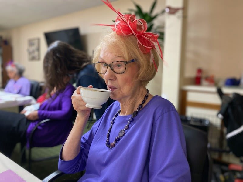 A woman wearing a purple outfit and a red hat enjoys a cup of tea indoors.