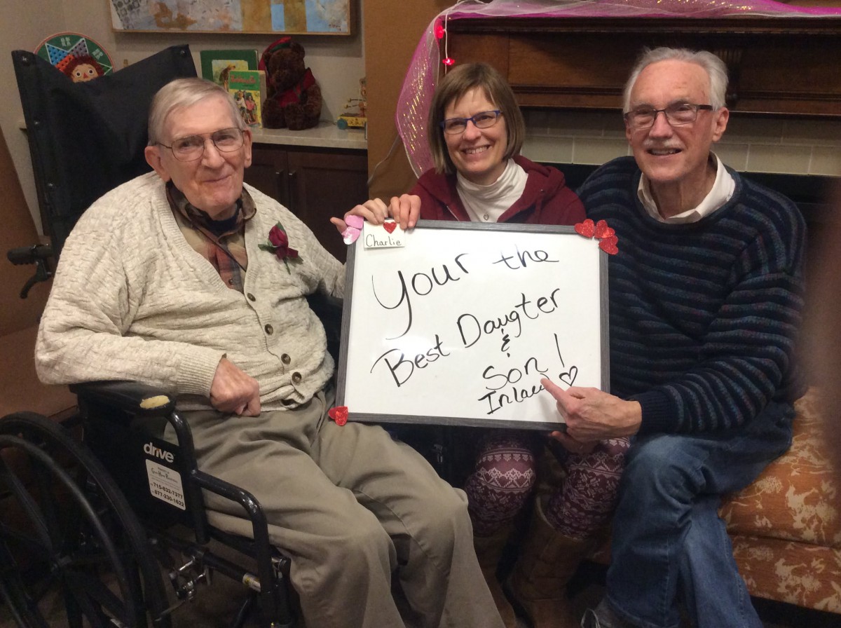 A group of three people, including an elderly man in a wheelchair, holding a sign that reads 'You're the best daughter' with smiling faces, gathered together indoors.