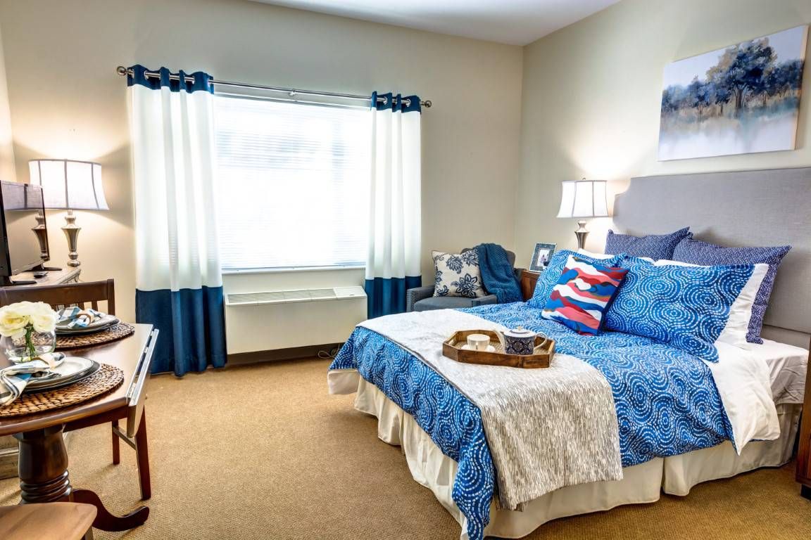 A cozy bedroom with a blue and white color scheme, featuring a neatly made bed, decorative pillows, a bedside lamp, and curtains framing the window.