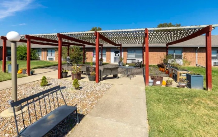 A sunny outdoor garden area featuring a pergola, benches, and a stone pathway.