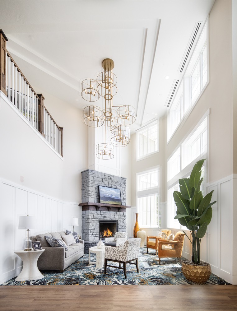 A spacious living room with high ceilings, featuring a modern chandelier, stone fireplace, and large windows that allow for ample natural light.