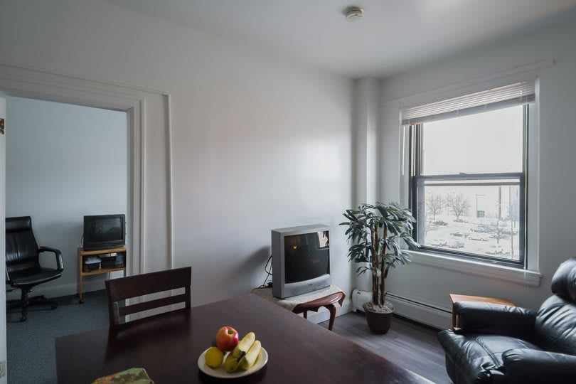 A cozy dining area with a table, fruit bowl, and a television, warmly lit by natural light from a large window.