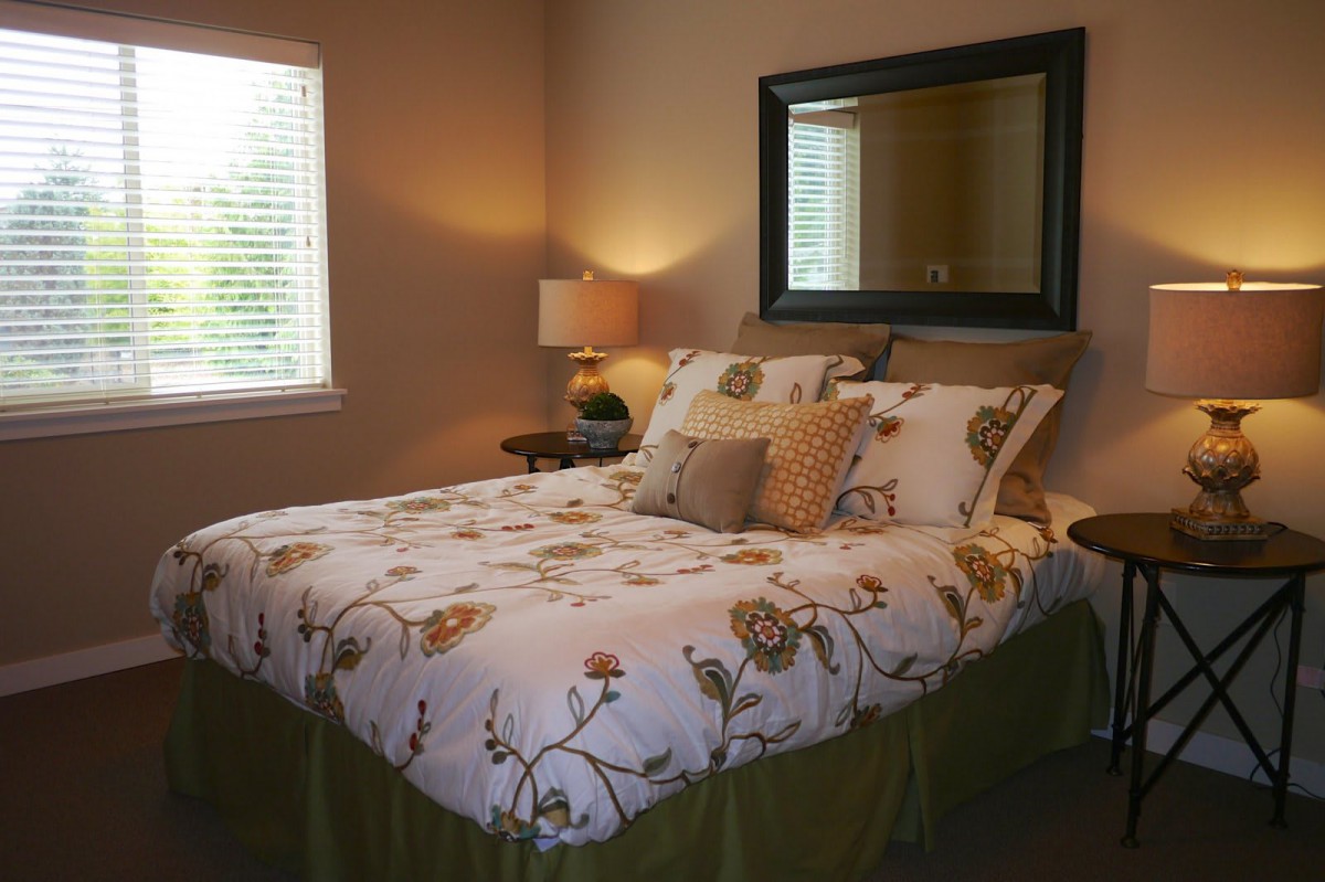 A cozy bedroom with floral bedding, a lamp on the nightstand, and a large mirror above the bed.