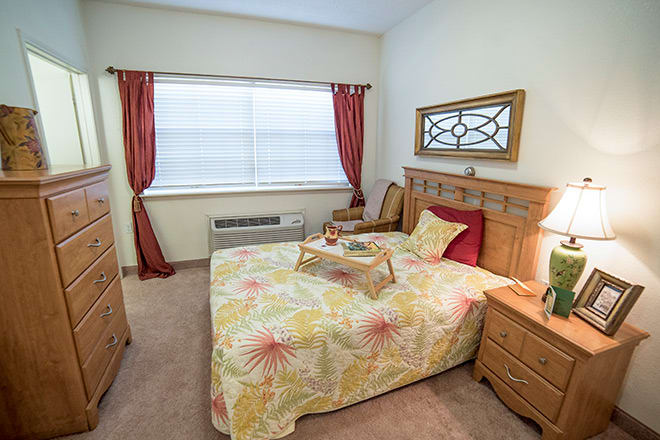 A cozy bedroom with a neatly made bed, decorative pillows, and a tray on the bed. The room features wooden furniture, accent curtains, and a large window covered by blinds.