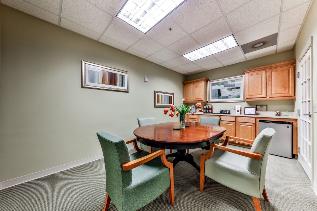 A small office meeting room with a round wooden table and several chairs, featuring artwork on the walls and a kitchenette in the background.