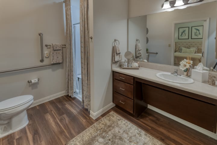 A modern bathroom with a wooden floor, featuring a sink and vanity area, a mirror, towels, and a walk-in shower.