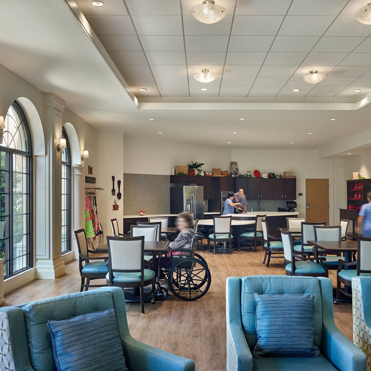 A spacious living area with comfortable seating, tables, and a kitchen in the background, featuring large windows and modern lighting.