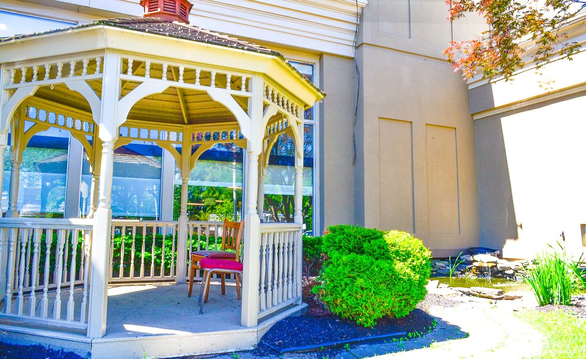 A charming gazebo with a wooden chair is surrounded by lush greenery on a sunny day.