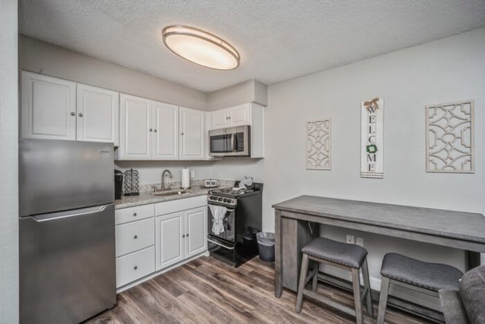 A modern kitchen with stainless steel appliances, white cabinets, and wood flooring. A small table with two chairs is set up in the corner.