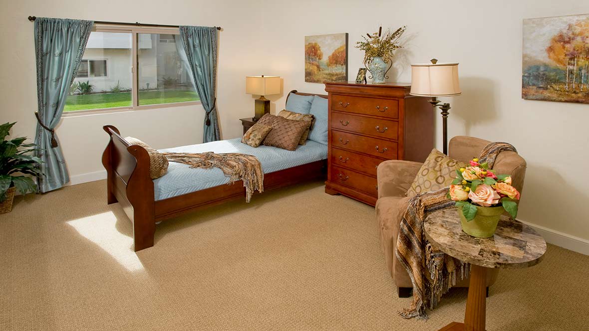 A cozy bedroom featuring a single bed, wooden dresser, and an armchair with decorations.