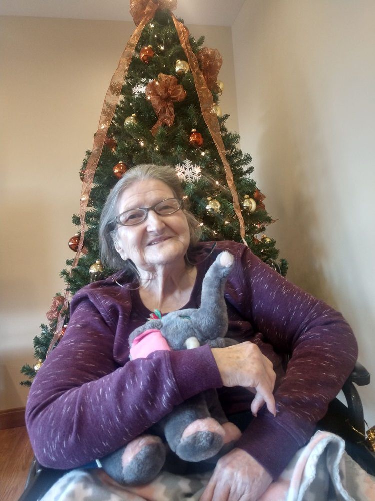 An elderly woman sitting in front of a Christmas tree, holding a stuffed animal.