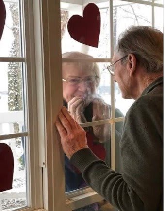 An elderly couple shares a tender moment through a window, illustrating connection despite separation.