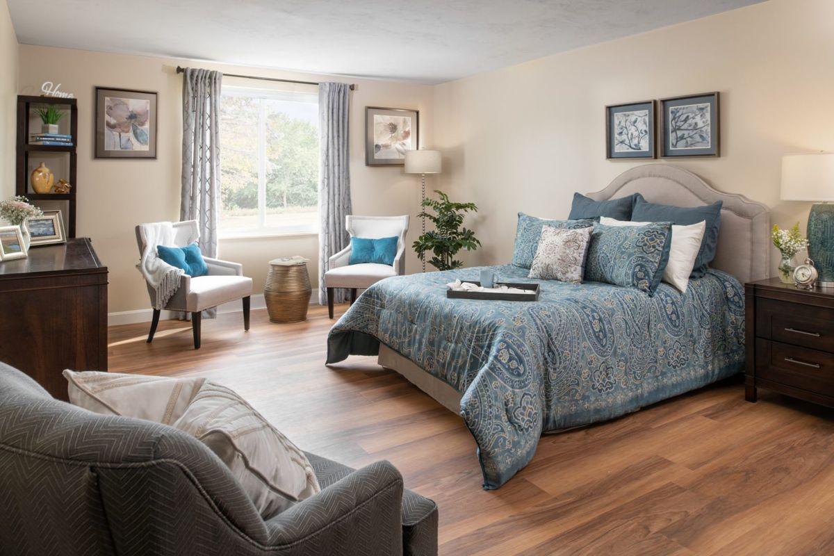 A cozy and elegantly designed bedroom with blue bedding, two chairs by the window, and soft lighting.