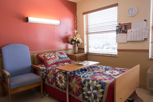 A cozy hospital patient room featuring a bed with a quilt, a chair, and floral decorations by the window.