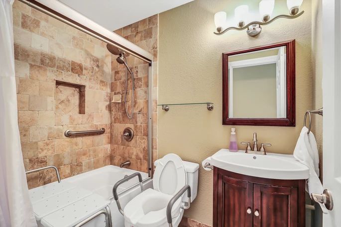 A cozy bathroom featuring a bathtub with a tile backsplash, a toilet equipped with handrails, and a sink with a mirror above it.