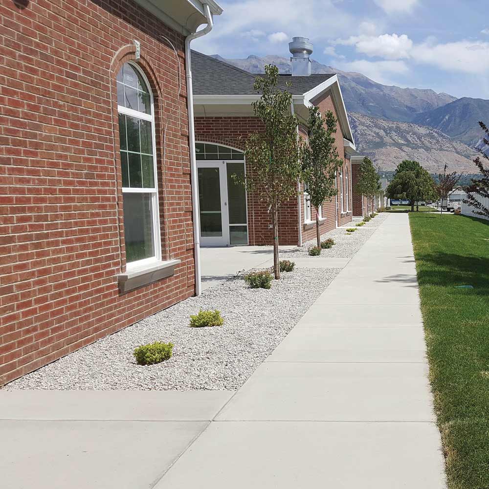 A brick building with large windows, bordered by a sidewalk and landscaped area with small plants, set against a backdrop of mountains and blue sky.