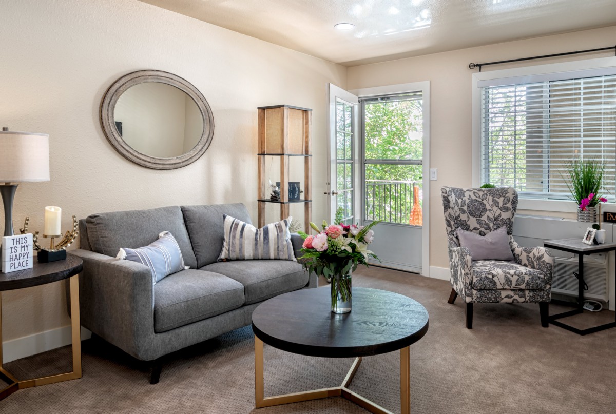 A cozy living room with modern decor, featuring a grey sofa, armchair, and a round coffee table adorned with a floral arrangement. A decorative mirror hangs on the wall, complementing the neutral color palette.