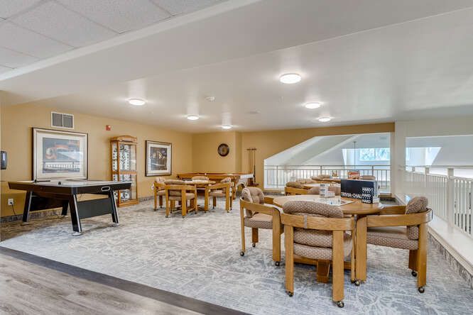 A spacious and well-lit community living area with wooden tables and chairs arranged on a carpeted floor.