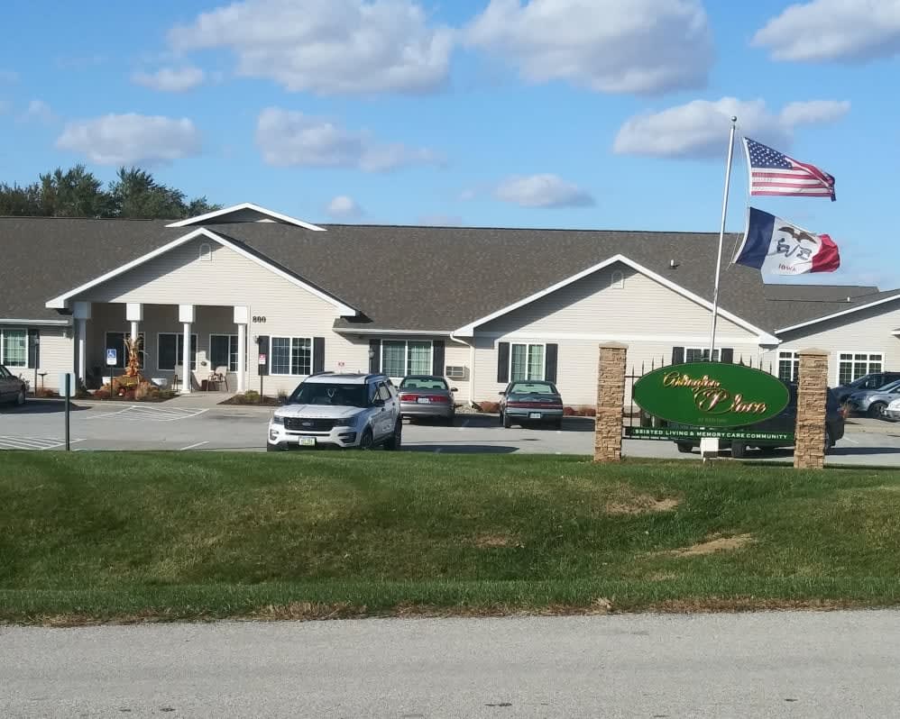 A single-story building with flags in front, surrounded by a lawn and parked cars under a partly cloudy sky.