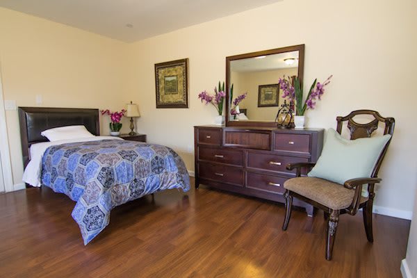 A cozy bedroom with a single bed, wooden dresser and mirror, decorated with flowers and wall art.