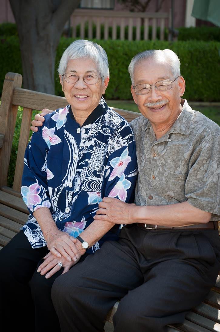 An elderly couple sitting on a bench, smiling warmly at the camera.