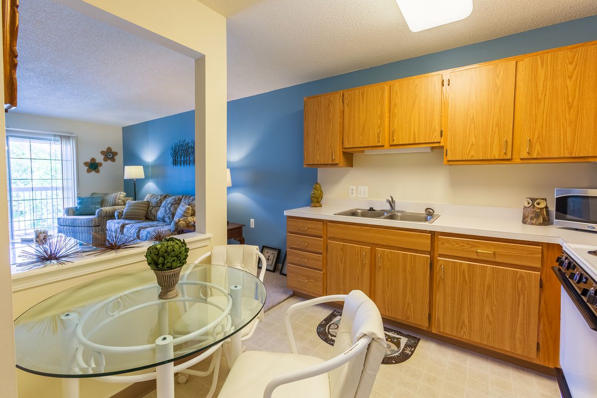 A cozy and modern open-concept apartment featuring a kitchen with wooden cabinets, an adjacent dining area with a glass table, and a living room in the background.