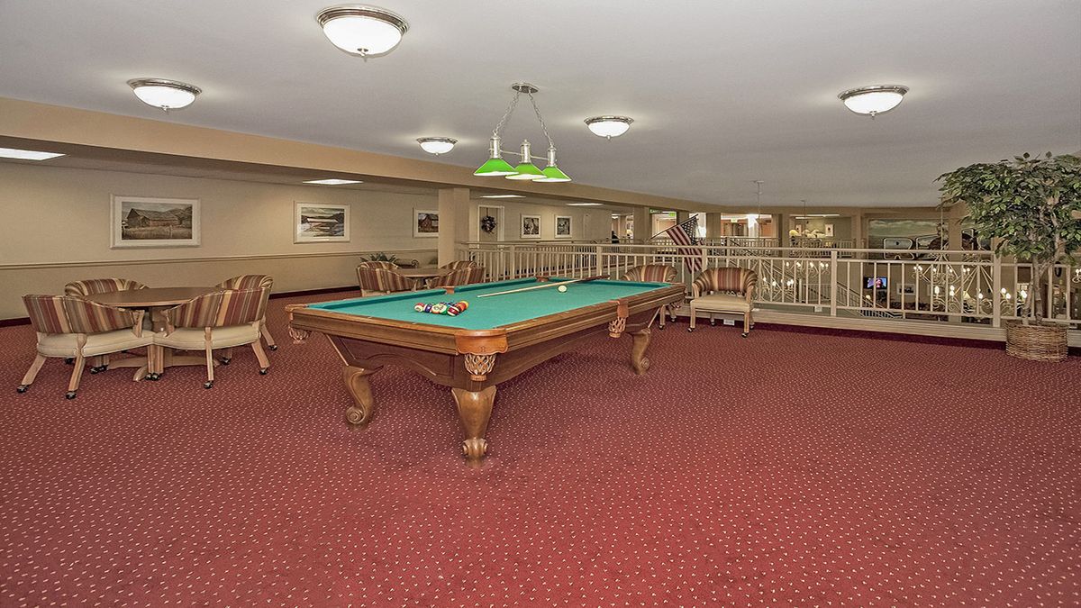 A spacious game room featuring a billiard table on a red carpeted floor, with overhead lighting and seating areas.