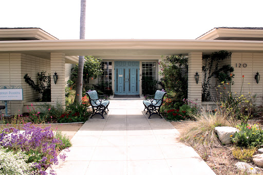 A charming house entrance with a blue door, surrounded by lush landscaping and comfortable outdoor seating.