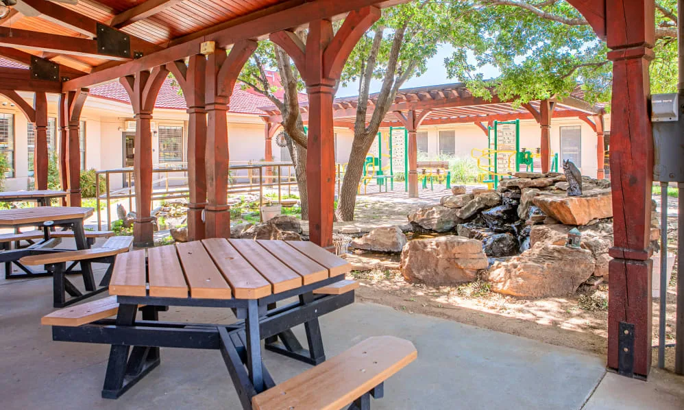 A serene outdoor setting featuring a wooden gazebo with a picnic table, surrounded by trees and rocks.