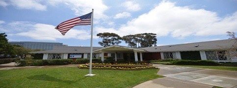 A building with an American flag in the foreground, surrounded by neatly arranged landscaping and trees.