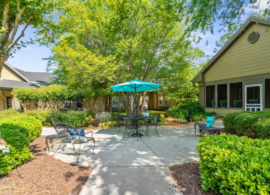 A serene outdoor patio area with chairs and a table under an umbrella, surrounded by lush greenery.