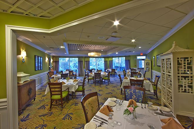 A cozy and well-lit dining room with neatly arranged tables and chairs, featuring green walls and a carpeted floor.