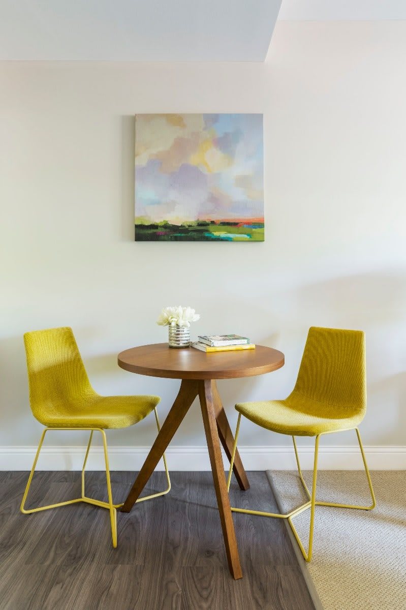 A modern dining area featuring two yellow chairs and a round wooden table, complemented by a colorful painting on the wall.